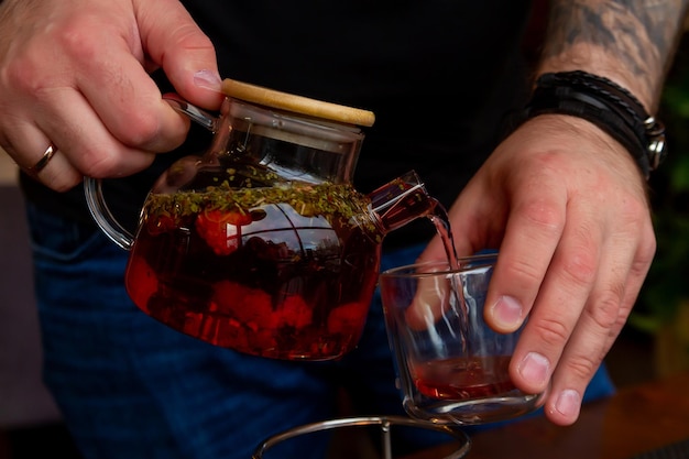 les mains des hommes versent le thé d'une théière en verre transparent dans une tasse transparente de tisane rouge