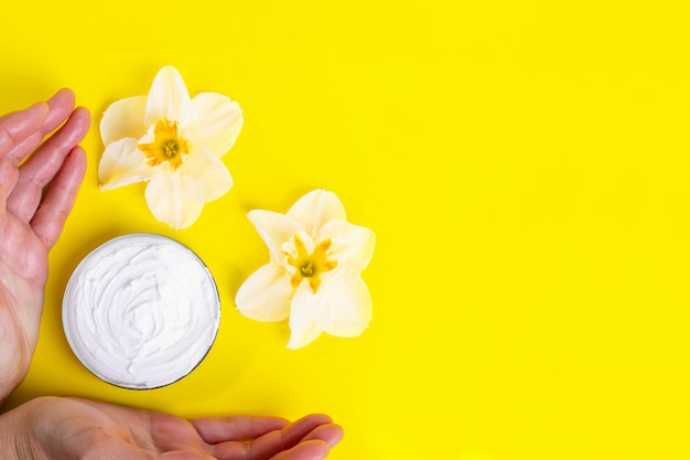 Des mains humaines prennent de la crème cosmétique en pot et de fleurs épanouies sur fond jaune vif.