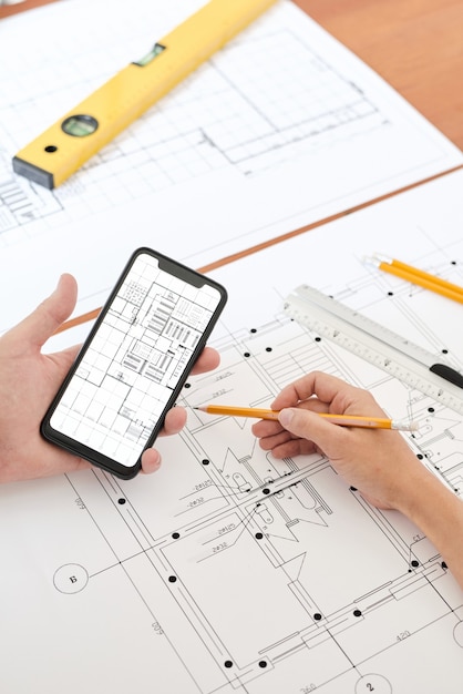 Photo mains d'un ingénieur ou d'un architecte professionnel moderne avec un crayon sur un croquis du nouveau bâtiment à l'aide d'un smartphone pour corriger ou vérifier les détails