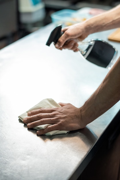 Mains d'un jeune chef contemporain de restaurant pulvérisant un désinfectant sur la table de la cuisine et la nettoyant avec un chiffon avant de préparer la nourriture