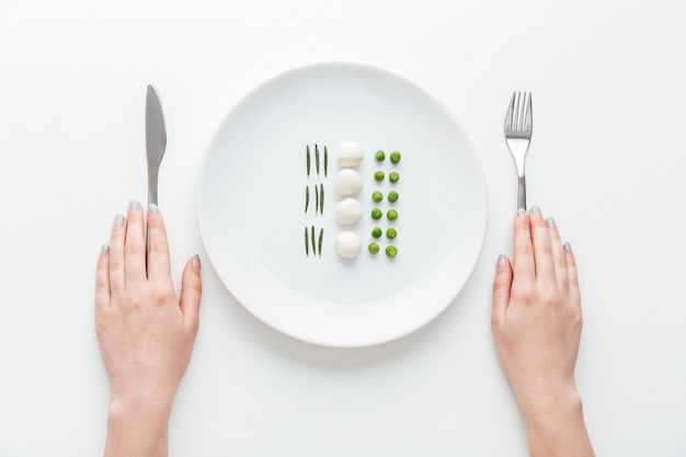 Mains de jeune femme avec couteau et fourchette mangeant des pois verts, de la mozzarella et des herbes sur la plaque