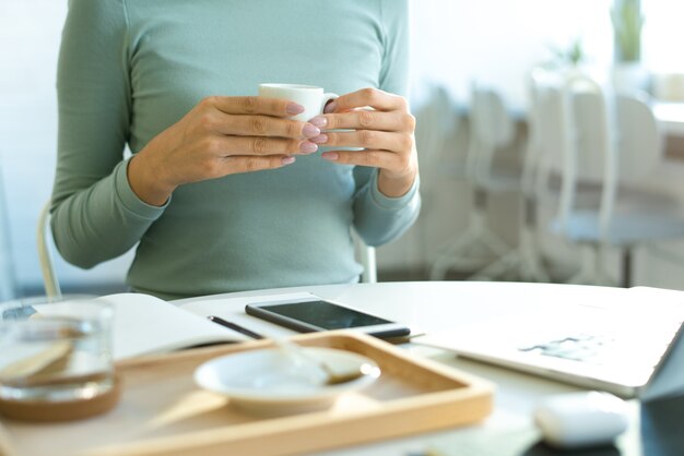 Mains de jeune femme décontractée tenant une tasse de thé sur la table avec des gadgets et un cahier ouvert tout en ayant une pause au café