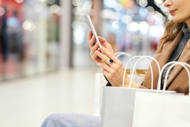 Mains de jeune femme élégante en tenue décontractée à la recherche d'annonces dans le smartphone tout en se reposant après le shopping dans le centre commercial