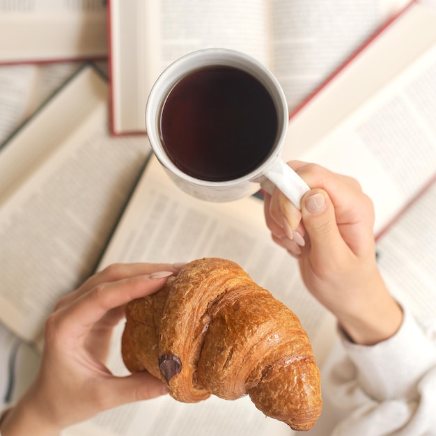 Mains d'une jeune femme mangeant un croissant avec du café sur le fond d'un tas de livres. Étudiant prenant son petit déjeuner, concept d'étude.