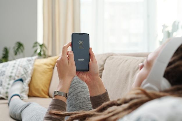 Mains de jeune femme reposante tenant le smartphone en face d'elle-même en position couchée sur le canapé et à la recherche de quelque chose à écouter