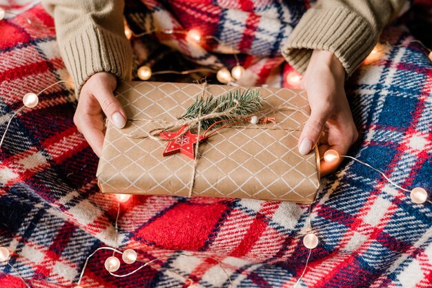 Mains de jeune femme tenant un coffret cadeau enveloppé avec étoile rouge et conifère sur le dessus sur plaid en laine à carreaux et guirlandes scintillantes