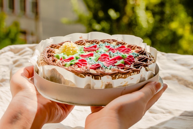 Photo mains de jeune femme tenant le gâteau aux noix rond traditionnel fraîchement ouvert de kiev dans l'emballage. les produits de boulangerie ukrainiens connus. journée ensoleillée et fond de verdure.