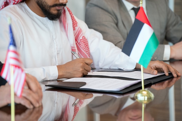 Photo mains d'un jeune homme d'affaires arabe ou d'un délégué en vêtements nationaux signant un contrat de partenariat commercial entre son pays et les états-unis