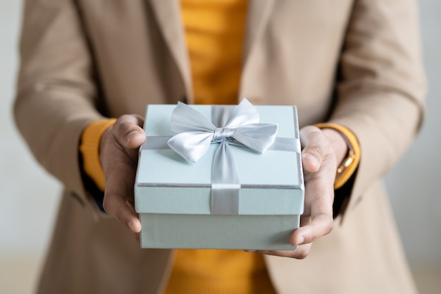 Mains de jeune homme d'affaires élégant dans des vêtements décontractés intelligents vous passant un cadeau emballé dans une boîte cadeau bleu clair avec un arc composé de ruban de soie sur le dessus