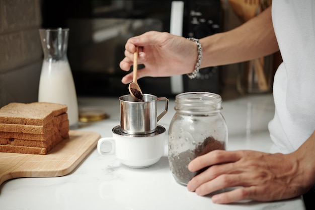 Mains de jeune homme mettant une cuillère de café moulu dans un goutteur en aluminium sur la table de la cuisine