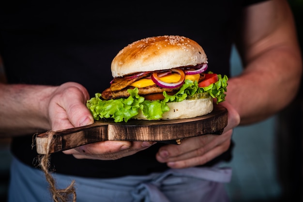 Mains mâles tenant un cheeseburger savoureux juteux avec du boeuf, de la laitue, des cornichons, des tomates et des rondelles d'oignon sur une table en bois. Cuisine de rue classique - burger grillé