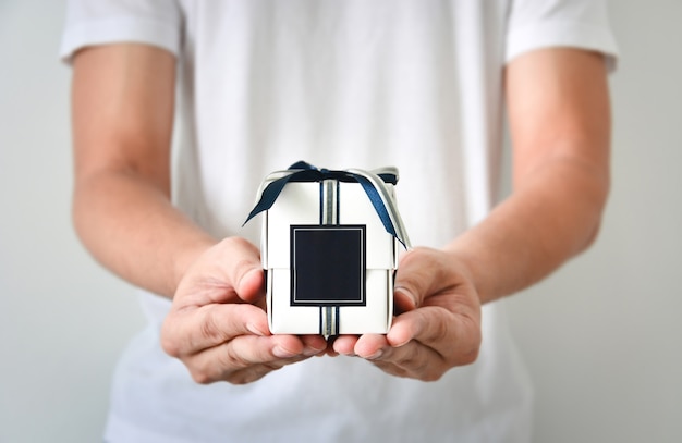 Mains mâles tenant une petite boîte-cadeau blanche enveloppée d'un ruban de couleur bleu et argent et d'une étiquette vierge bleu foncé