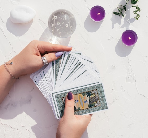 Des mains avec des ongles et des anneaux violets tiennent un jeu de cartes de tarot sur une surface blanche. Boule de cristal, bougies et pierres, vue de dessus