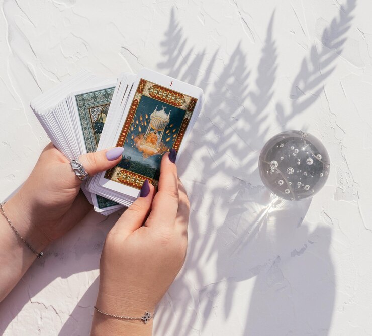 Des mains avec des ongles et des anneaux violets tiennent un jeu de cartes de tarot sur une surface blanche. Boule de cristal et ombre de feuille de fougère, vue de dessus