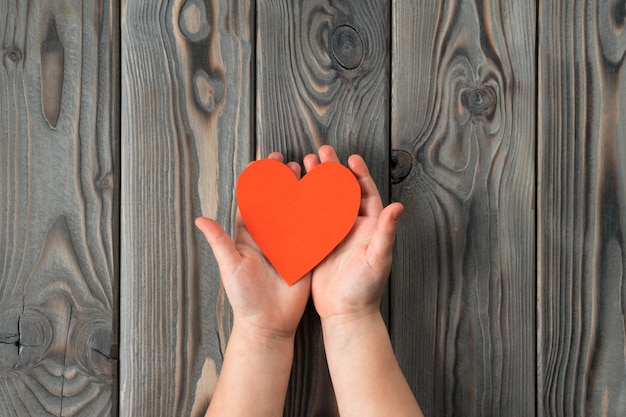 Des mains ouvertes méconnaissables tiennent et montrent un petit coeur en papier rouge sur fond de bois Famille et enfants ou Saint Valentin