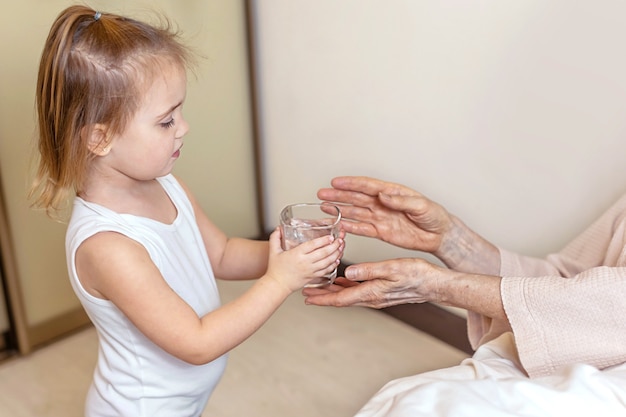 Les mains d'une petite fille donnent un verre d'eau à une grand-mère âgée dans le lit à la maison. Le concept de prise en charge des adultes, de soutien, d'amour, d'assistance, d'entraide