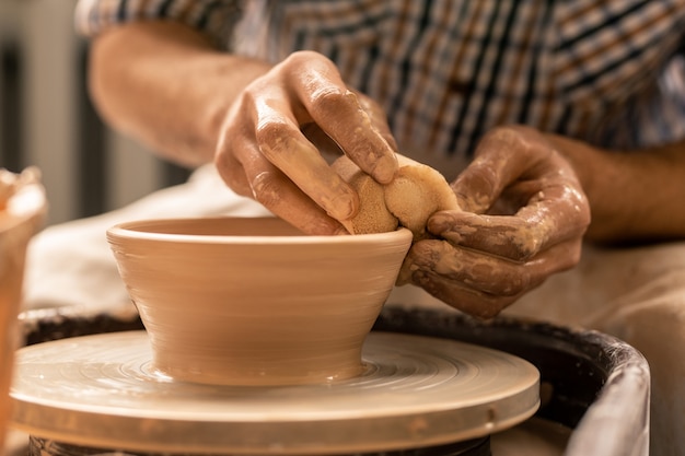 Mains de potier prenant l'excès d'argile sur le bord du pot brut tout en travaillant en tournant le tour de poterie