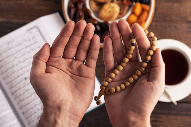 Mains de prière avec chapelet en bois sur fond de livre Coran, tasse de thé, assiette de fruits secs, concept iftar, mois de Ramadan, vue de dessus, gros plan