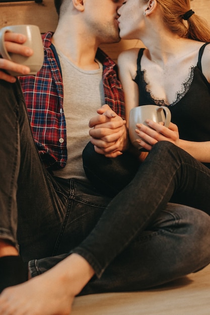 Mains avec des tasses de café chaud d'un charmant jeune couple assis sur le sol dans la cuisine s'embrassant le matin.