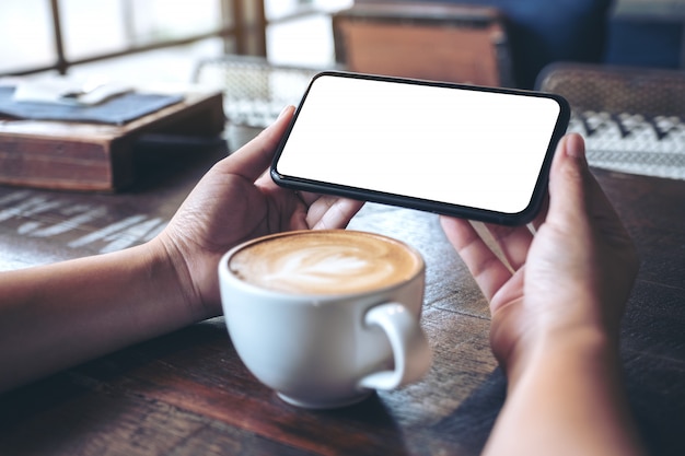 Mains tenant et à l'aide d'un téléphone mobile noir avec écran blanc horizontalement pour regarder avec une tasse de café sur une table en bois