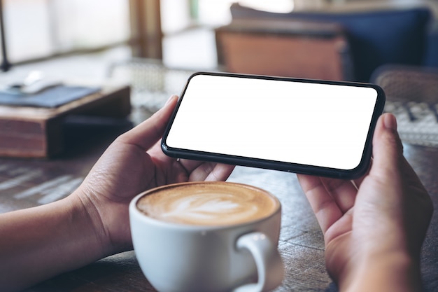 Mains Tenant Et à L'aide D'un Téléphone Mobile Noir Avec écran Blanc Horizontalement Pour Regarder Avec Une Tasse De Café Sur Une Table En Bois