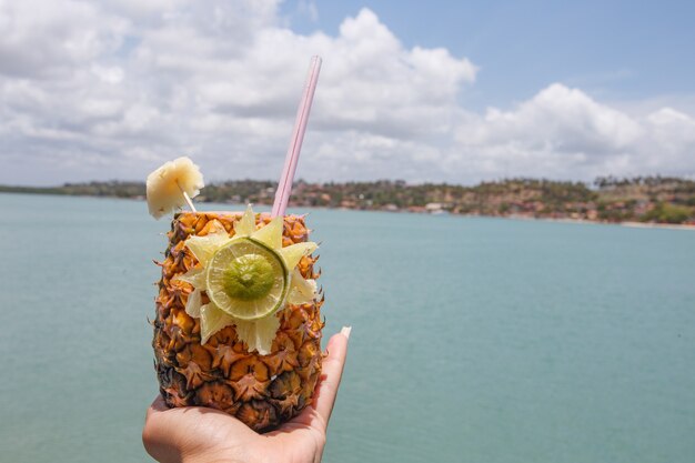 Mains tenant une boisson à l'ananas avec fond de plage.