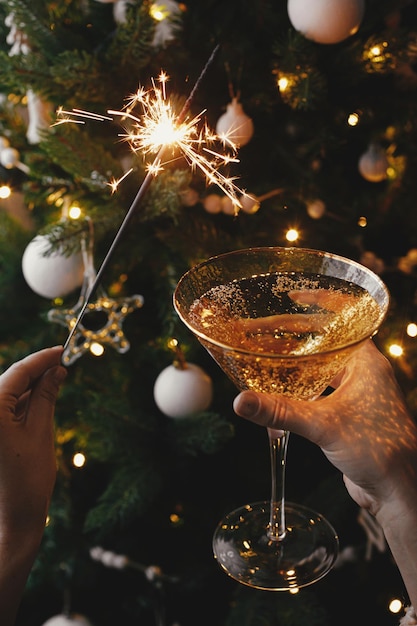 Photo mains tenant un cierge magique brûlant et un verre de champagne contre les lumières de l'arbre de noël bonne année