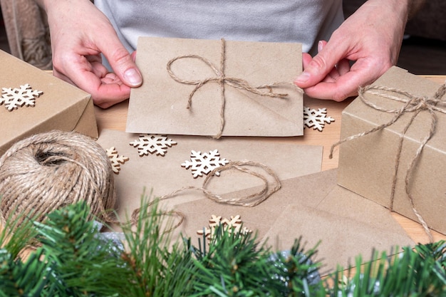 Mains tenant une enveloppe en papier kraft avec une lettre de Noël sur fond de cadeaux de Noël, de papier d'emballage, de ficelle et de flocons de neige. Concept de style écologique de Noël. Concept zéro déchet.