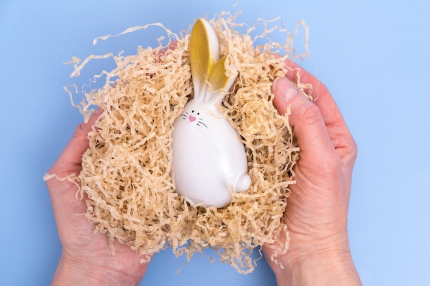 Mains tenant un lapin de Pâques blanc assis dans un nid fait maison sur fond bleu Composition créative minimale de Pâques