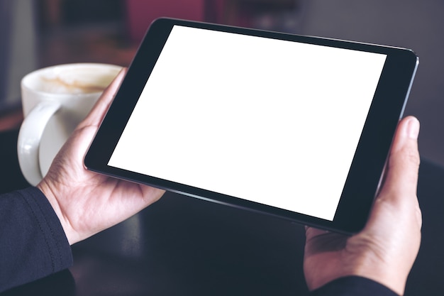 mains tenant une tablette noire avec un écran blanc et une tasse de café sur fond de table