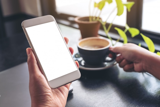 Mains tenant un téléphone mobile blanc avec écran blanc tout en buvant du café