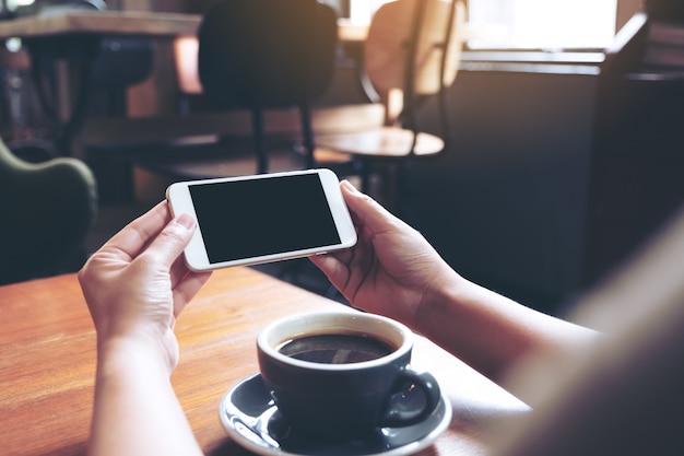 mains tenant et utilisant un téléphone mobile blanc avec écran noir blanc horizontalement pour regarder avec une tasse de café sur une table en bois