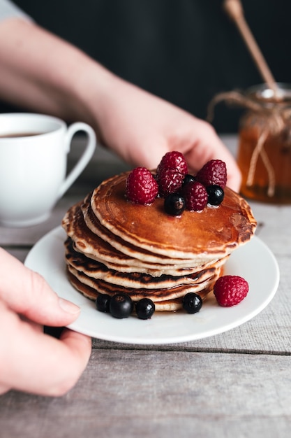 Mains tiennent une assiette blanche avec des crêpes, des baies, du miel, une tasse de café sur une table en bois, un pot et une cuillère. Photo de haute qualité