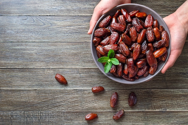 Les mains tiennent des dattes séchées dans un bol en céramique sur une table en bois. Fruits secs bio. Vue de dessus. Mise à plat avec espace de copie