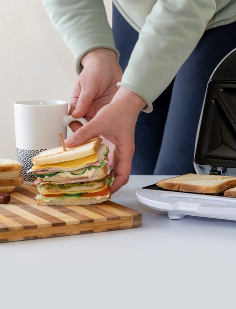 Les mains tiennent un sandwich frais cuit avec du fromage, du bacon et des légumes près du toster et une tasse de café