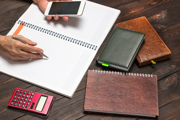 Les mains tiennent le stylo et le smartphone, le cahier ouvert. Sur la calculatrice de table et les cahiers. Vue de dessus.