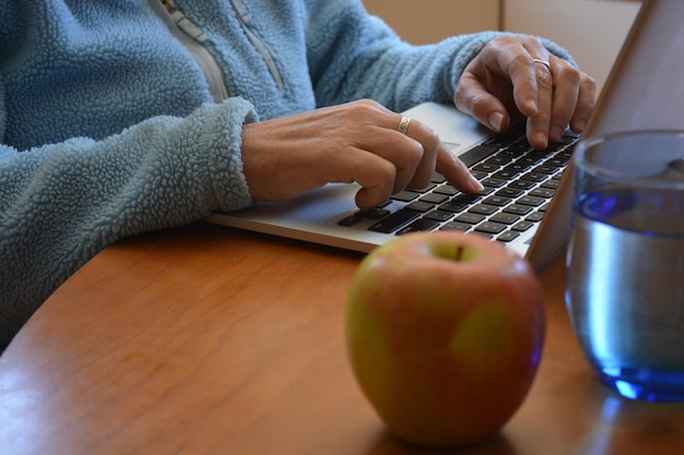 Photo des mains en train de taper sur le clavier de l'ordinateur portable à angle bas avec une pomme et un verre d'eau au premier plan