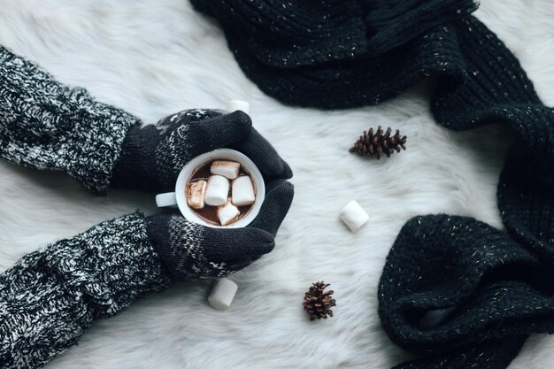 Les mains utilisant des vêtements et des gants d'hiver chauds tiennent une tasse de chocolat chaud sur un tapis de fourrure blanche