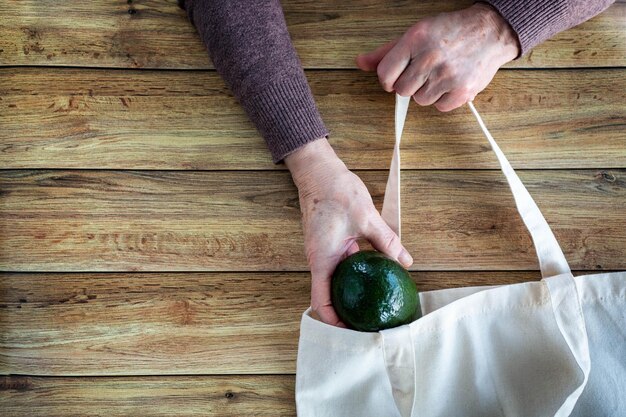 Les mains d'une vieille femme mettent de l'avocat biologique frais dans un sac à provisions écologique sur fond de bois Mise à plat