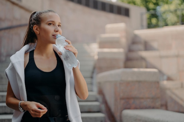 Maintenir l'équilibre hydrique Une jeune sportive épuisée boit une boisson rafraîchissante après l'entraînement prend une pause porte un t-shirt et un pull blanc autour des épaules a soif Concept sport et santé