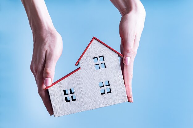 Maison En Bois Blanche Dans Les Mains D'une Femme. Fond Bleu. Concept De Logement