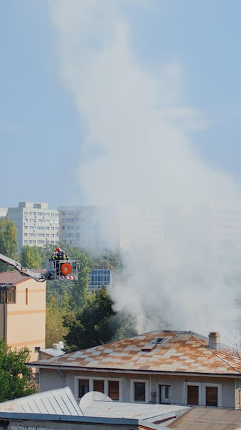 Maison brûlant dans le quartier de la ville et pompiers aidant avec un camion plate-forme et de l'eau pour éteindre le feu. Pompiers essayant d'éteindre les flammes chaudes et la fumée sur le toit du bâtiment.