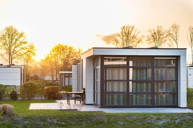 Maison de campagne moderne avec une grande pelouse et une clôture en bois Devant la maison il y a une terrasse couverte avec un coin salon Sur la pelouse il y a des arbres et des parterres de fleurs Il fait soleil Dehors