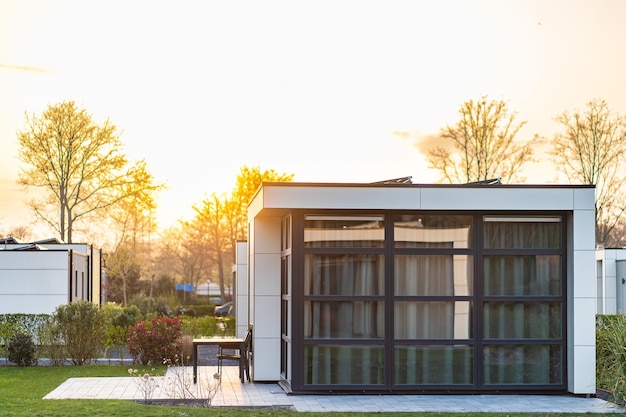 Maison de campagne moderne avec une grande pelouse et une clôture en bois Devant la maison il y a une terrasse couverte avec un coin salon Sur la pelouse il y a des arbres et des parterres de fleurs Il fait soleil Dehors