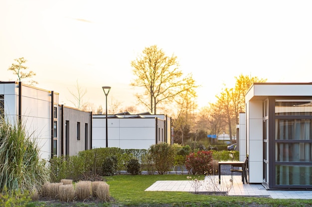 Maison de campagne moderne avec une grande pelouse et une clôture en bois Devant la maison il y a une terrasse couverte avec un coin salon Sur la pelouse il y a des arbres et des parterres de fleurs Il fait soleil Dehors