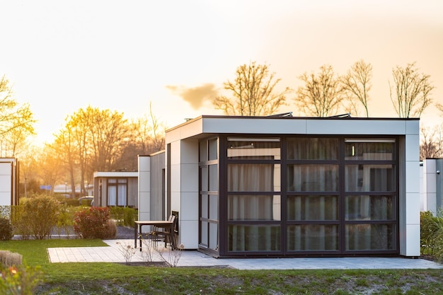 Maison de campagne moderne avec une grande pelouse et une clôture en bois Devant la maison il y a une terrasse couverte avec un coin salon Sur la pelouse il y a des arbres et des parterres de fleurs Il fait soleil Dehors