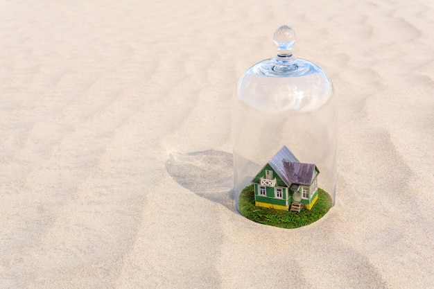 Maison en carton jouet avec pelouse verte protégée par une cloche en verre au milieu d'un désert de sable sans vie