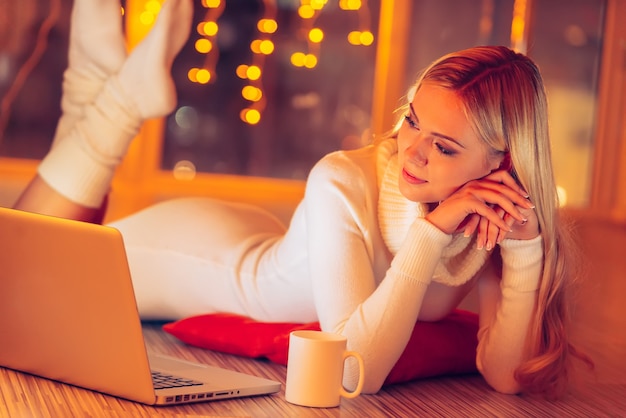 La Maison Est Le Meilleur Endroit Au Monde. Belle Jeune Femme En Pull Blanc Et Chaussettes Allongé Sur Le Sol Et Regardant Un Ordinateur Portable Avec Des Lumières De Noël En Arrière-plan