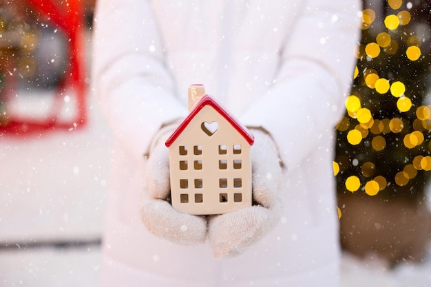 Maison miniature cottage entre les mains d'une femme portant des mitaines et des vêtements chauds en plein air dans la neige Affaire pour l'achat immobilier construction relocalisation hypothèque Maison confortable Noël nouvel an réservation