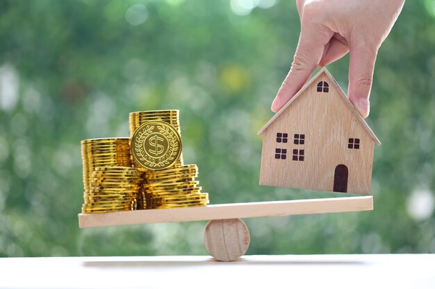 Maison modèle avec une pile de pièces d'or, de l'argent sur une balance en bois, une balançoire avec un fond vert naturel, un concept d'investissement commercial et immobilier.